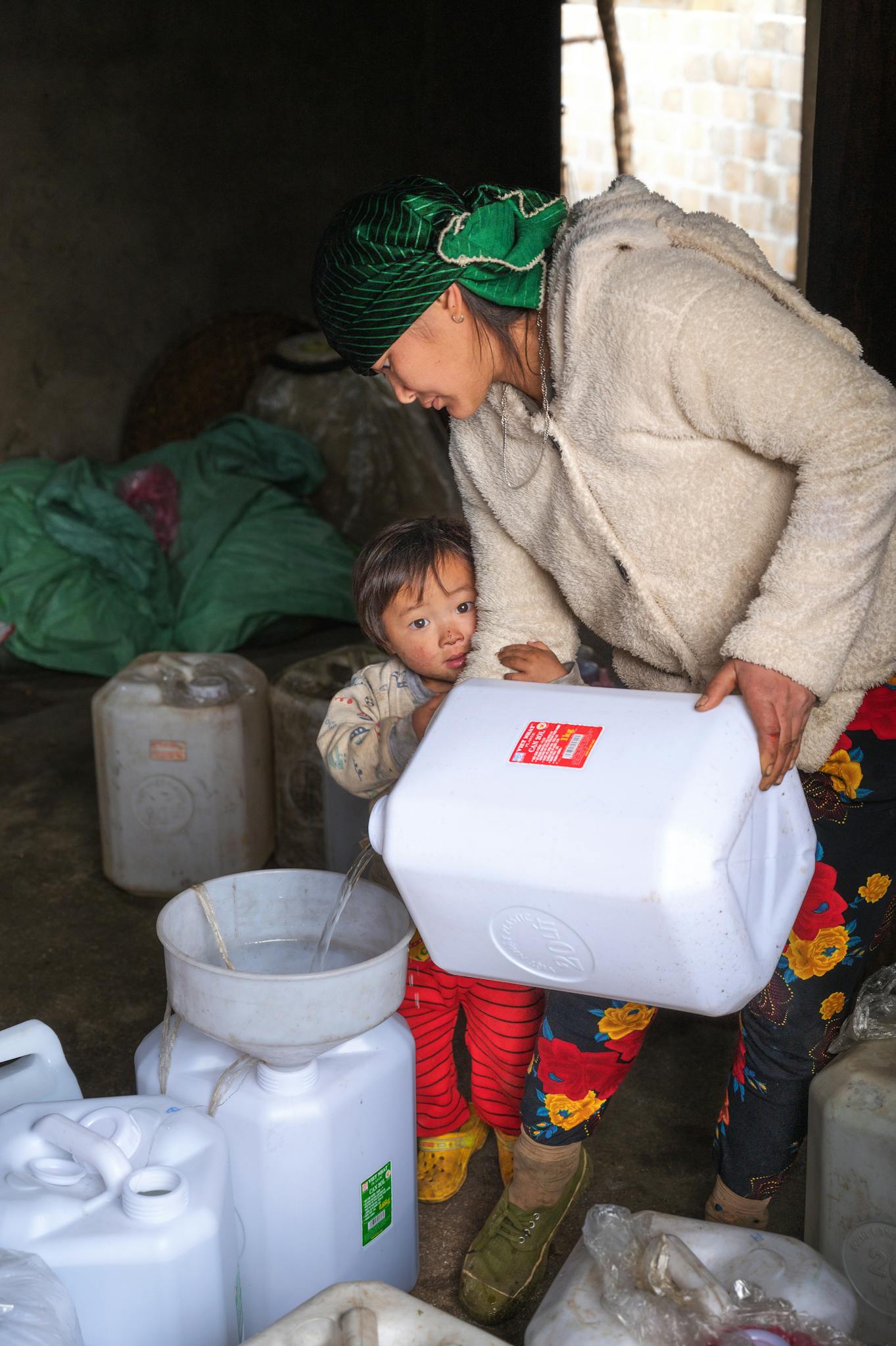 A woman and child pour water from a large container indoors, focused on their task.