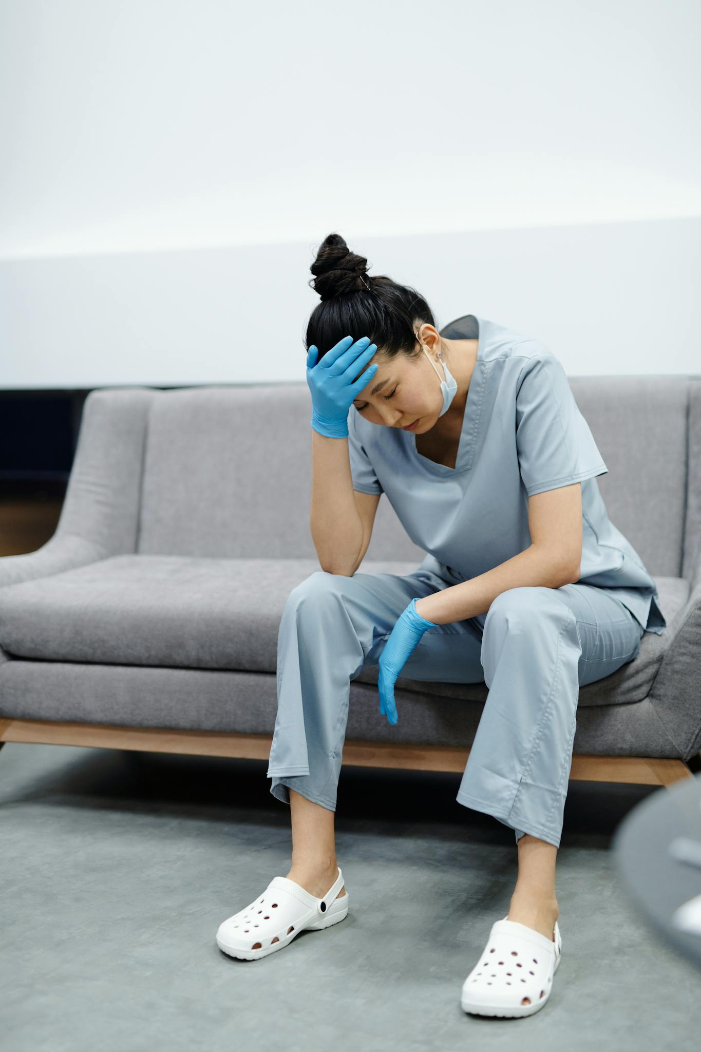 Tired nurse in scrubs sitting on a sofa, looking exhausted with her hand on her head.