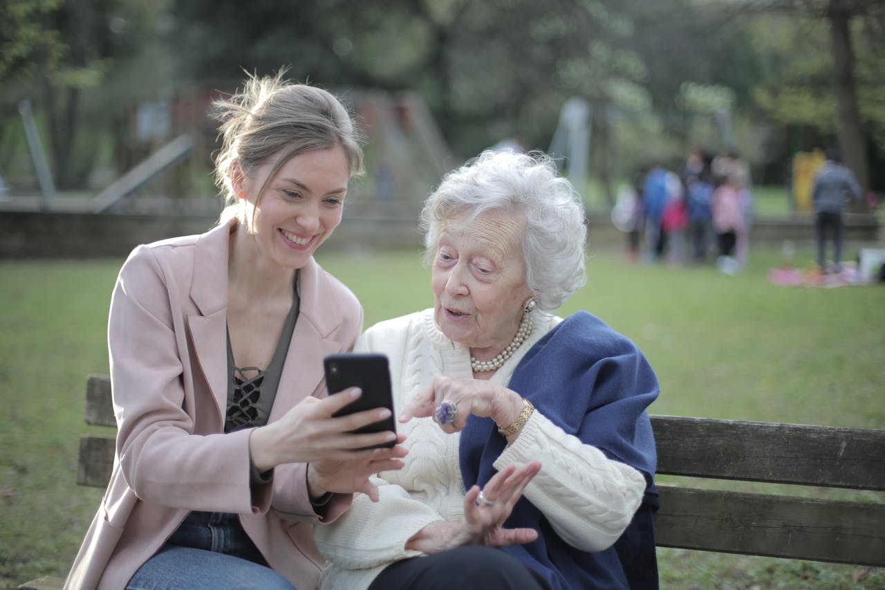 Delighted female relatives sitting together on wooden bench in park and browsing mobile phone while learning using